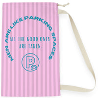 This laundry bag features a pink background with vertical pastel-pink stripes. Centered, bold, teal text reads: "All the good ones are taken." Above the text, in a large curved arc, it says: “Men are like parking spaces!” Below the quote is a parking symbol: a capital letter “P” inside a circle with a car icon in the corner. The bag includes a drawstring closure and a canvas carrying strap. The tone is playful, retro, and full of tongue-in-cheek charm. It is the small version.