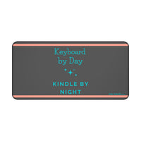 A charcoal gray desk mat features a playful message split across two lines: "Keyboard by Day" in turquoise serif text on the left, and "Kindle by Night" in sans-serif turquoise on the right. Turquoise starburst-style sparkles accent the design, and two thin orange horizontal lines run along the top and bottom edges. This is the largeversion.