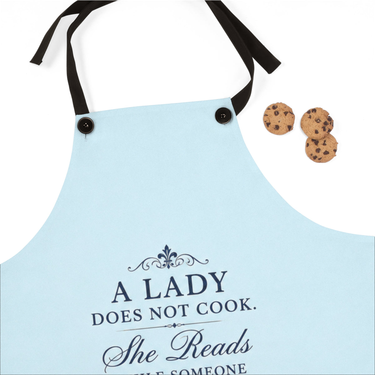 A light blue kitchen apron with black neck and waist straps, featuring the printed text 'A LADY DOES NOT COOK. She Reads WHILE SOMEONE' in dark navy lettering.