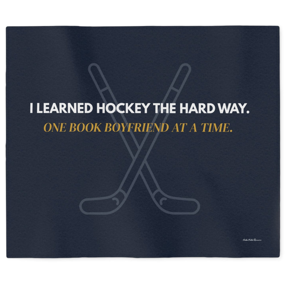 A  dark navy sweatshirt blanket featuring crossed hockey sticks and the text 'I LEARNED HOCKEY THE HARD WAY.' in white and 'ONE BOOK BOYFRIEND AT A TIME in gold.