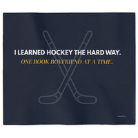 A  dark navy sweatshirt blanket featuring crossed hockey sticks and the text 'I LEARNED HOCKEY THE HARD WAY.' in white and 'ONE BOOK BOYFRIEND AT A TIME in gold.