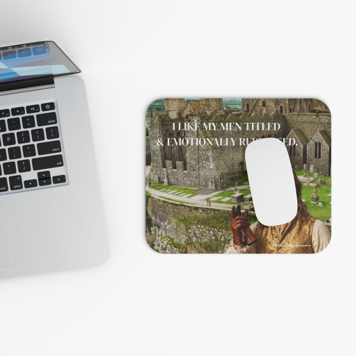 Regency romance mouse pad featuring a handsome aristocratic man in period clothing before a castle with the text “I Like My Men Titled & Emotionally Repressed." adjacent to a laptop with a white computer mouse on the mouse pad.