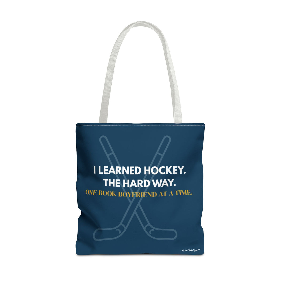 Navy blue hockey romance tote bag with white handles, crossed hockey sticks and the words “I Learned Hockey. The Hard Way” in white and "One Book Boyfriend at a Time" in gold.