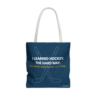 Navy blue hockey romance tote bag with white handles, crossed hockey sticks and the words “I Learned Hockey. The Hard Way” in white and "One Book Boyfriend at a Time" in gold.