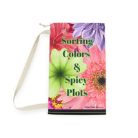 This laundry bag features a vivid collage of oversized watercolor-style flowers, including pink zinnias, orange gerberas, red hibiscus, a purple dahlia, and a bright green orchid. Centered over the floral background is bold black text with a lime green shadow that reads: “Sorting Colors &Spicy Plots”. At the bottom of the bag is a black bar with a thin lime green line in the middle. The bag includes a drawstring closure and sturdy canvas carrying strap. This is the large bag.