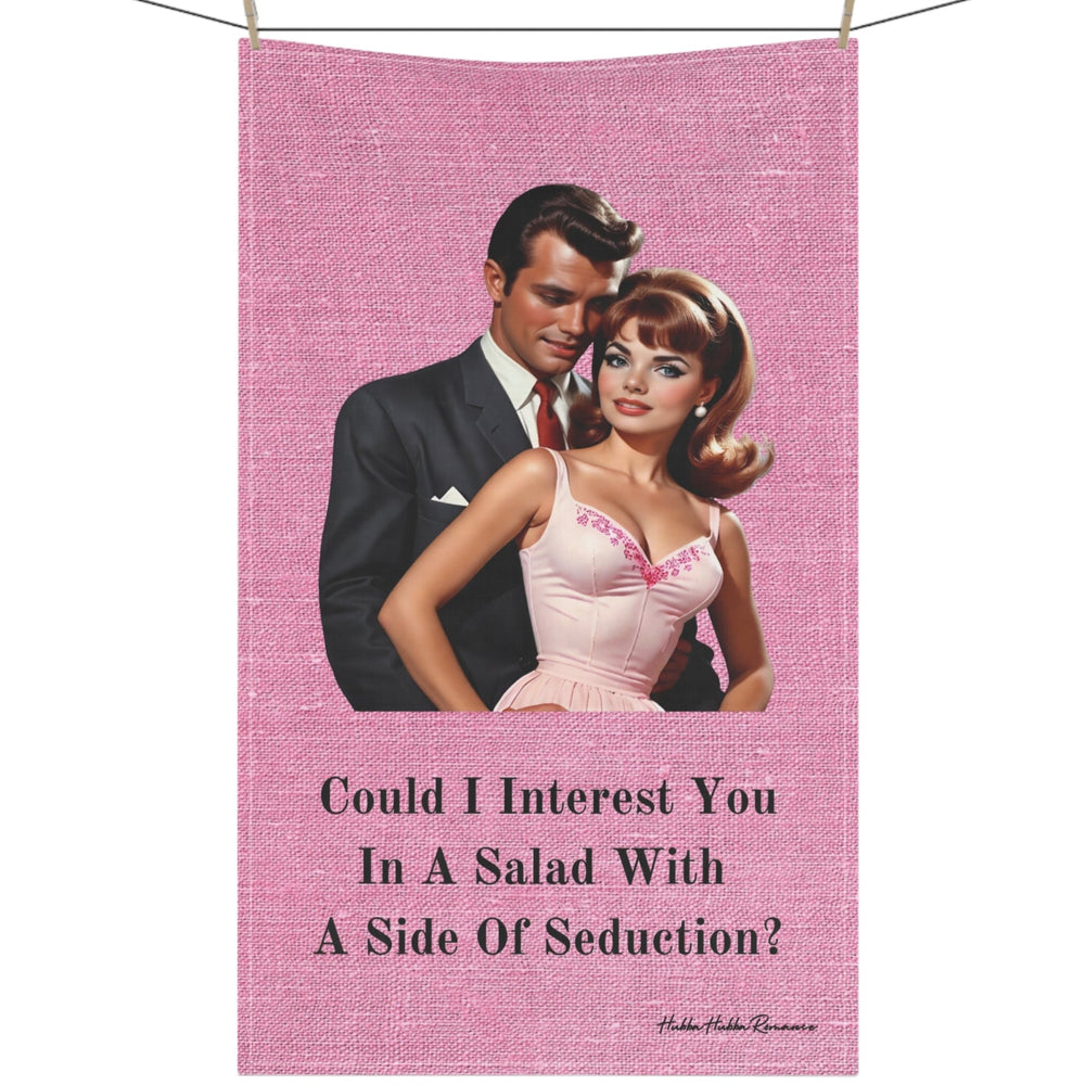 The image is a vintage-style illustration of a sophisticated couple from the 1950s. The woman, styled with glamorous makeup and a bouffant hairstyle, wears a pink dress with floral embroidery, while the man, exuding charm, is dressed in a sleek, dark suit and stands close behind her. Above them, text reads: "Could I Interest You In A Salad With A Side Of Seduction?" This design blends retro fashion with a playful, romantic flair, adding a touch of nostalgic wit to a kitchen and is displayed on a clothesline