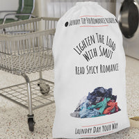 This laundry bag features a white background with a colorful pile of clothes at the bottom. Above the pile, black text reads: “Lighten the Load with Smut” in an arched layout. Below that, in a straight line, it says: “Read Spicy Romance.” At the top, the header “Laundry Tip for Romance Readers” is centered between two black dots and underlined in red. The bottom of the image reads: “Laundry Day Your Way” in black text, also underlined in red. The bag includes a drawstring closure and a canvas carry strap.