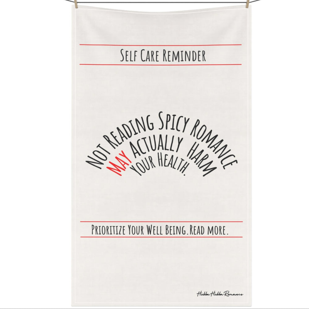 An off white cotton kitchen towel that is text based with the following message displayed in black text: "Self-Care Reminder” in a banner format at the top of the towel. Under it is “Not Reading Spicy Romance May Actually Harm Your Health” in 3 levels of nested arched text. “Prioritize Your Well-being. Read More." Is also in a banner format under the arched text. The saying is meant to be sarcastically ironic. The towel is displayed on a clothesline.