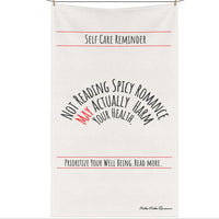 An off white cotton kitchen towel that is text based with the following message displayed in black text: "Self-Care Reminder” in a banner format at the top of the towel. Under it is “Not Reading Spicy Romance May Actually Harm Your Health” in 3 levels of nested arched text. “Prioritize Your Well-being. Read More." Is also in a banner format under the arched text. The saying is meant to be sarcastically ironic. The towel is displayed on a clothesline.