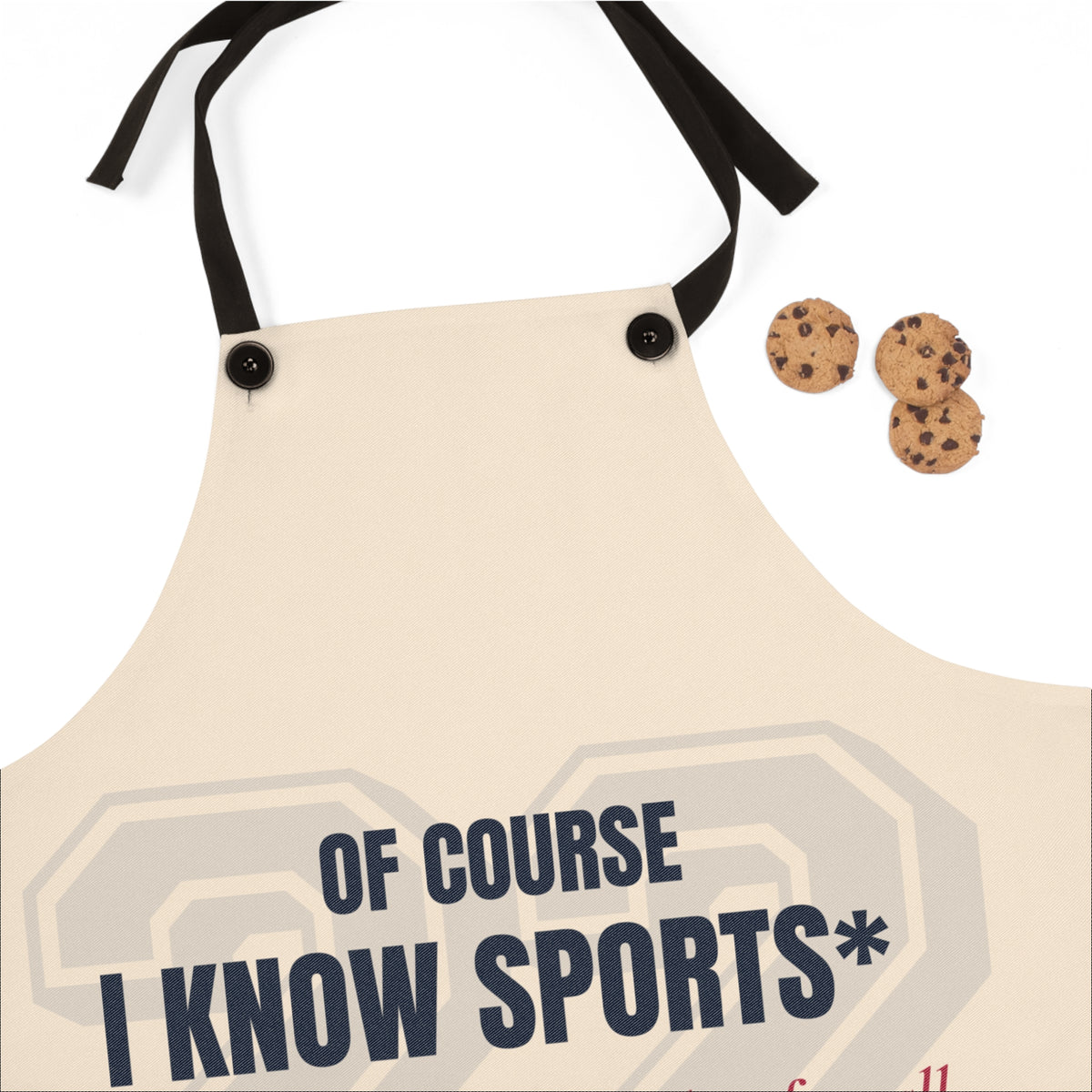 A beige apron with black neck and waist straps, featuring the text 'OF COURSE I KNOW SPORTS*' printed in dark navy blue. Three chocolate chip cookies are placed to the right of the apron on a clean white background.