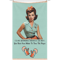 The kitchen towel features a vintage-inspired illustration of a glamorous woman from the 1950s, sporting a stylish updo and a mint green camp shirt, black belt, and an orange skirt, accompanied by text that reads, "I Like Romance Novels So Hot You Need Oven Mitts to Turn the Pages." Below the text are two whimsical oven mitts with playful cats and orange polka-dot details. The design combines retro charm and humor, appealing to fans of steamy romance novels. The napkin is displayed on a clothesline.