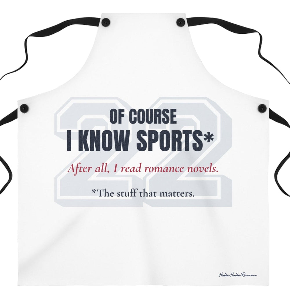 A white bib-style apron with black neck and waist ties, featuring a sports-themed graphic print that reads 'OF COURSE I KNOW SPORTS*' and 'After all, I read romance novels.' on a plain white background. There is an asterisk qualifying the sports knowledge to the stuff that matters.