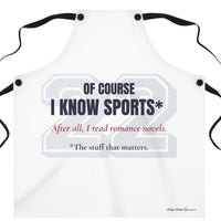 A white bib-style apron with black neck and waist ties, featuring a sports-themed graphic print that reads 'OF COURSE I KNOW SPORTS*' and 'After all, I read romance novels.' on a plain white background. There is an asterisk qualifying the sports knowledge to the stuff that matters.