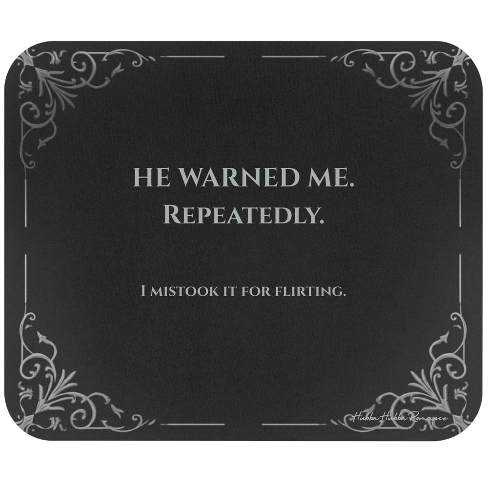 A rectangular black mouse pad with rounded corners, featuring light grey text and light gray filigree corner accents. The text  reads 'HE WARNED ME. REPEATEDLY. I mistook it for flirting.