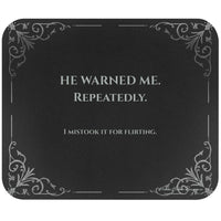 A rectangular black mouse pad with rounded corners, featuring light grey text and light gray filigree corner accents. The text  reads 'HE WARNED ME. REPEATEDLY. I mistook it for flirting.