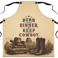 A beige apron with black neck and waist ties, featuring a western-themed graphic print with the text 'BURN THE DINNER. KEEP THE COWBOY.