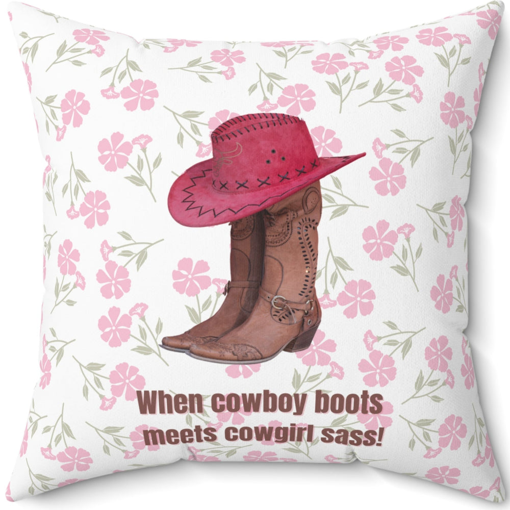 This 20 inch square pillow features a pair of brown leather cowboy boots with intricate stitching and metal accents, topped with a red cowboy hat with black stitched detailing. The background is soft white with a delicate pink floral pattern. Below the boots and hat, the bold brown text reads: "When cowboy boots meets cowgirl sass!" The design combines Western charm with a playful, feminine flair.