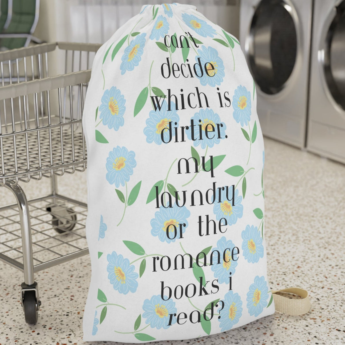 This design features light blue daisy-style flowers with yellow centers and green leaves on a white background. Black text reads: “Can’t decide which is dirtier: my laundry or the romance books I read?” for a cheeky, flirtatious message. The overall look is clean and pretty with a spicy twist for romance readers. There is a draw string enclosure and a large canvas carry strap. The bag is the larger size and is displayed in front of a cart for context.