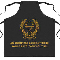 A black bib-style apron with gold printed graphics and text on the front, shown flat against a plain white background. The design features a gold chef's cloche and crossed utensils emblem with the text 'Est CHAPTER 3' inside a laurel wreath, and the phrase 'MY BILLIONAIRE BOOK BOYFRIEND WOULD HAVE PEOPLE FOR THIS.