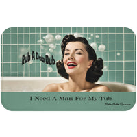 This bath mat features a retro-style image of a glamorous woman with styled dark hair, red lipstick, and a joyful smile, lounging in a bubble bath. Behind her is a sea-green tiled bathroom wall with soap bubbles floating around. Playful black text reads “Rub A Dub Dub.” At the bottom of the mat, centered over a seafoam green background strip, bold black text reads: “I Need A Man For My Tub.” The overall vibe is vintage pin-up with playful humor. This is the larger version.