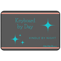 A charcoal gray desk mat features a playful message split across two lines: "Keyboard by Day" in turquoise serif text on the left, and "Kindle by Night" in sans-serif turquoise on the right. Turquoise starburst-style sparkles accent the design, and two thin orange horizontal lines run along the top and bottom edges.