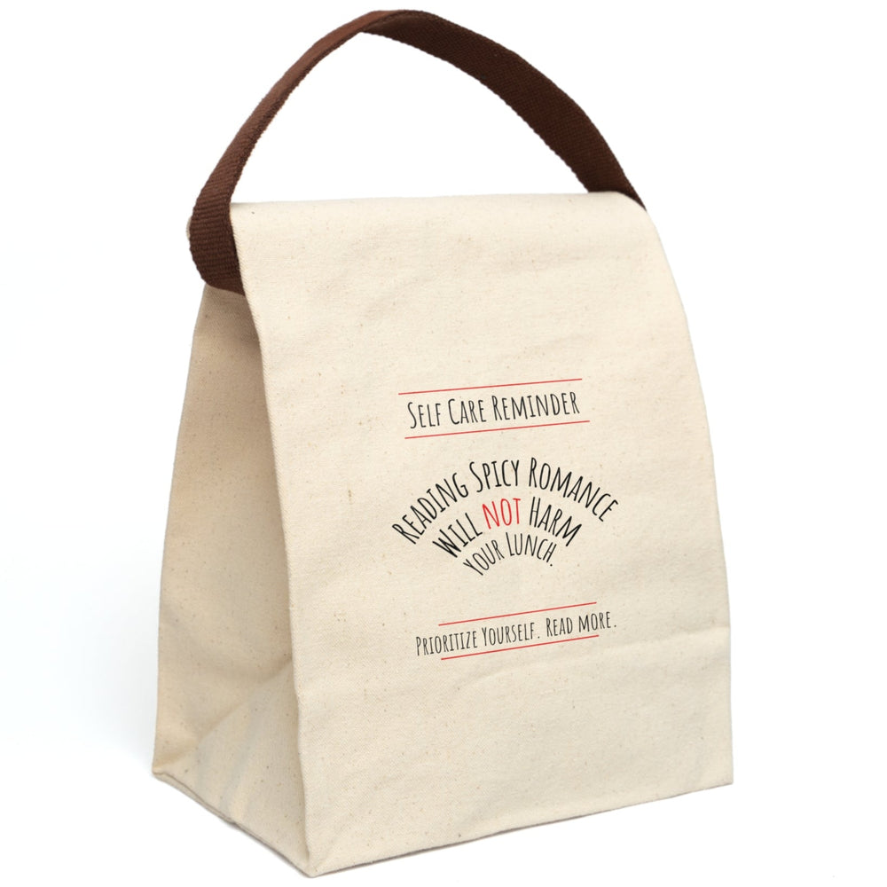 This canvas lunch bag design features black and red text on a white background. At the top, bold lettering reads: “Self-Care Reminder,” underscored with a red line. The main message is displayed in a curved arch and reads, “Reading Spicy Romance Will Not Harm Your Lunch,” with the word “Not” highlighted in red. At the bottom, text reads: “Prioritize Yourself. Read More,” also framed with red lines. The bag has a roll-top closure and a sturdy canvas carrying strap which is displayed upright.