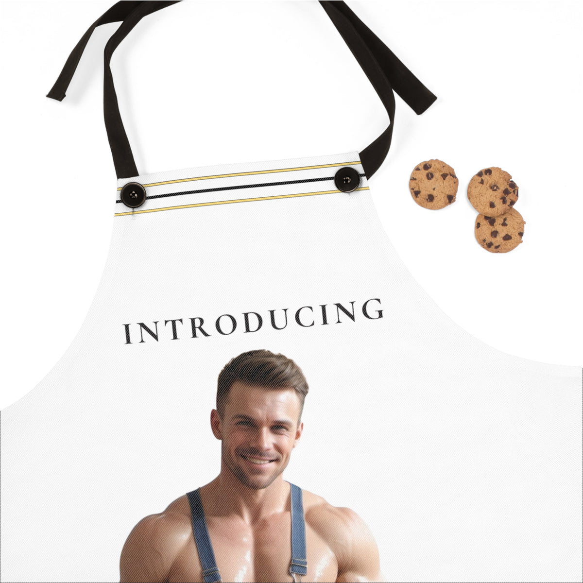 A white apron with black neck straps and a thin gold horizontal stripe, featuring the text 'INTRODUCING' and a printed photo of a shirtless muscular man wearing blue overalls. Three chocolate chip cookies are placed to the right on a white background.