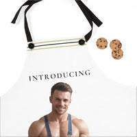 A white apron with black neck straps and a thin gold horizontal stripe, featuring the text 'INTRODUCING' and a printed photo of a shirtless muscular man wearing blue overalls. Three chocolate chip cookies are placed to the right on a white background.