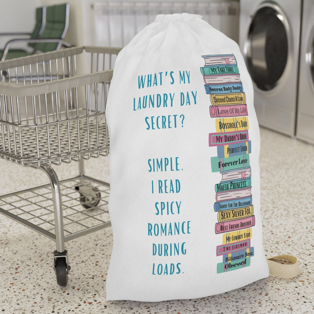This is an image of an off white laundry bag with a drawstring enclosure at the top and a large canvas carrying strap on the side. The bag has an image of a large stack of romance books with various spicy titles. Next to the stack is text that says: What's My Laundry Secret? Simple. I read Spicy Romance During Loads. This is the larger bag and is displayed in front of a laundry cart for size context.