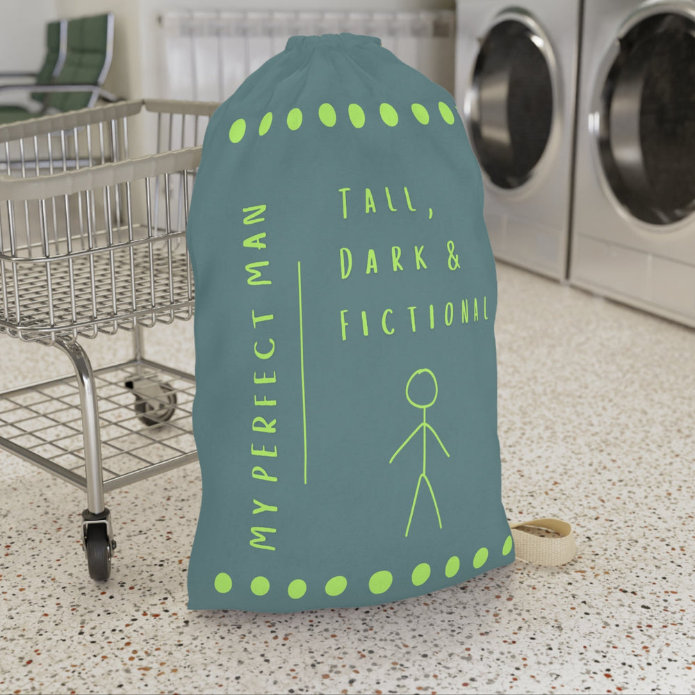This laundry bag features a minimalist design on a muted greenish-gray background. A lime green stick figure stands next to bold lime green text that reads “Tall, Dark & Fictional,” with the words “My Perfect Man” printed vertically along the side. Rows of lime-green dots border the top and bottom of the bag. The bag includes a durable canvas carrying strap and a drawstring enclosure for secure, easy transport. This is the large version. It is in front of a cart for sizing context.