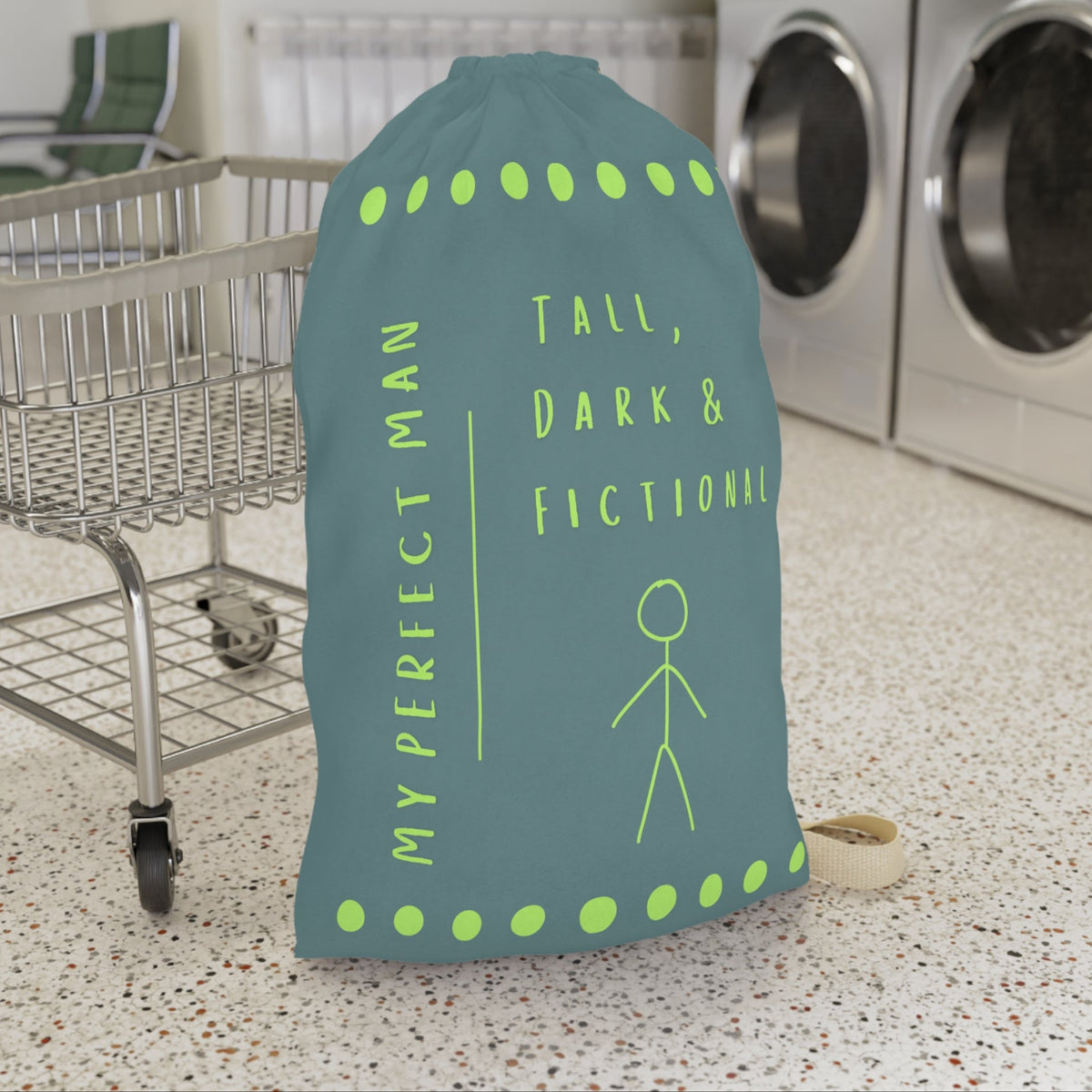 This laundry bag features a minimalist design on a muted greenish-gray background. A lime green stick figure stands next to bold lime green text that reads “Tall, Dark & Fictional,” with the words “My Perfect Man” printed vertically along the side. Rows of lime-green dots border the top and bottom of the bag. The bag includes a durable canvas carrying strap and a drawstring enclosure for secure, easy transport. This is the large version. It is in front of a cart for sizing context.