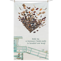 Hanging on a clothesline with clothespins is an oversized kitchen towel featuring a beach scene with a green lifeguard tower and a man looking up, along with tourists on the beach. In the sky, there is a swirling tornado of books. The text reads: Look Up. A Romance Book-Nado Is Headed Our Way. It is a tongue-in-cheek reference to all the beach books read by sun worshippers.