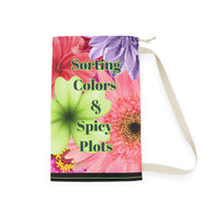 This laundry bag features a vivid collage of oversized watercolor-style flowers, including pink zinnias, orange gerberas, red hibiscus, a purple dahlia, and a bright green orchid. Centered over the floral background is bold black text with a lime green shadow that reads: “Sorting Colors &Spicy Plots”. At the bottom of the bag is a black bar with a thin lime green line in the middle. The bag includes a drawstring closure and sturdy canvas carrying strap. This is the large bag.