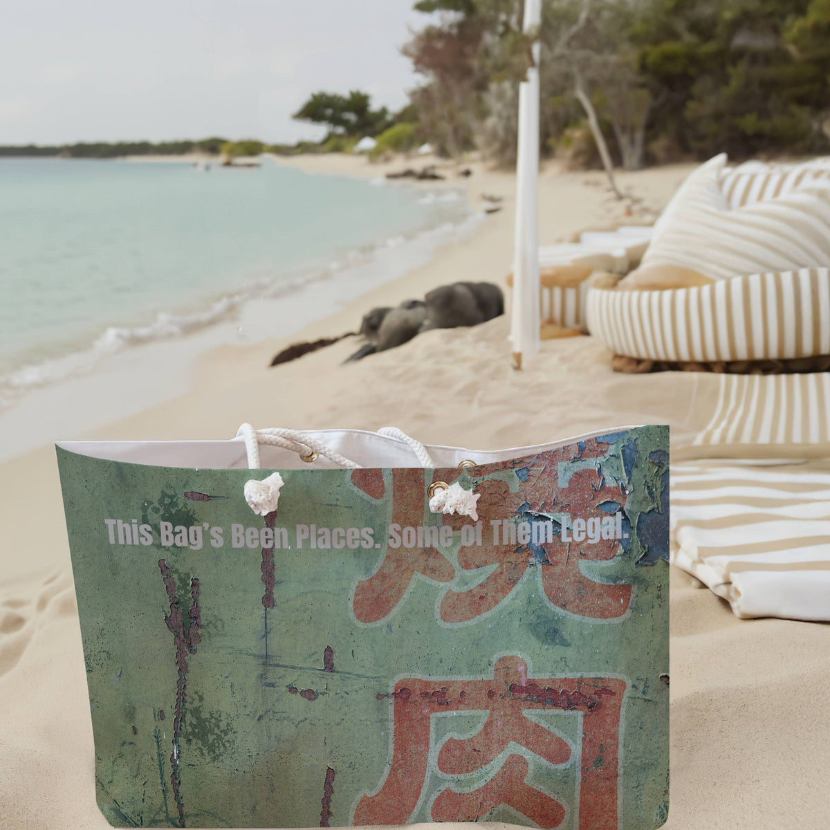 The weekender bag is displayed standing upright on the beach next to beach towels.