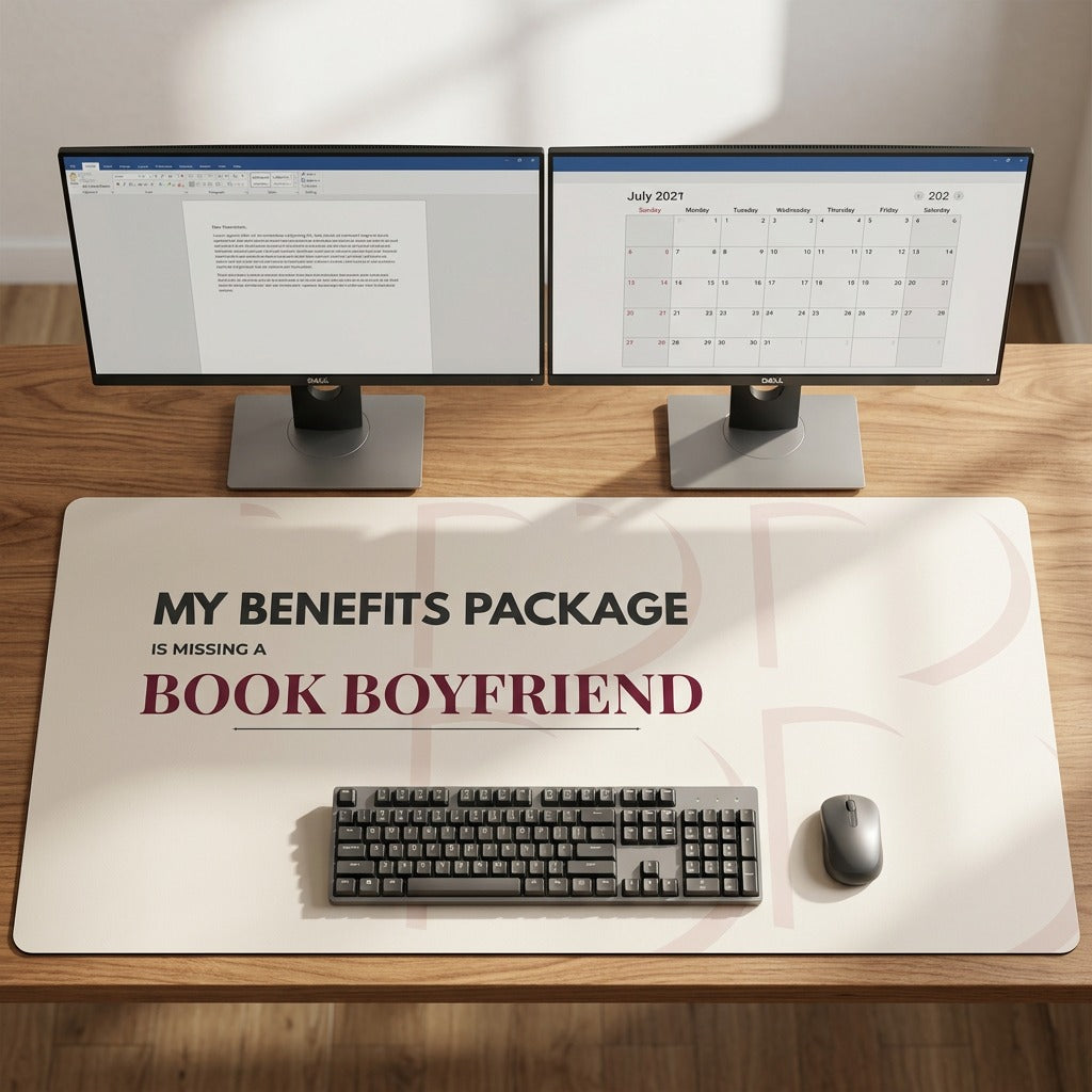 A large tan desk mat with the text 'MY BENEFITS PACKAGE IS MISSING A BOOK BOYFRIEND' in black and burgundy lettering, styled on a wooden desk under two computer monitors, a keyboard, and a mouse. This is the largest size offered.
