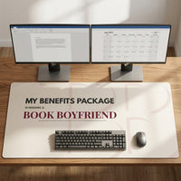 A large tan desk mat with the text 'MY BENEFITS PACKAGE IS MISSING A BOOK BOYFRIEND' in black and burgundy lettering, styled on a wooden desk under two computer monitors, a keyboard, and a mouse. This is the largest size offered.