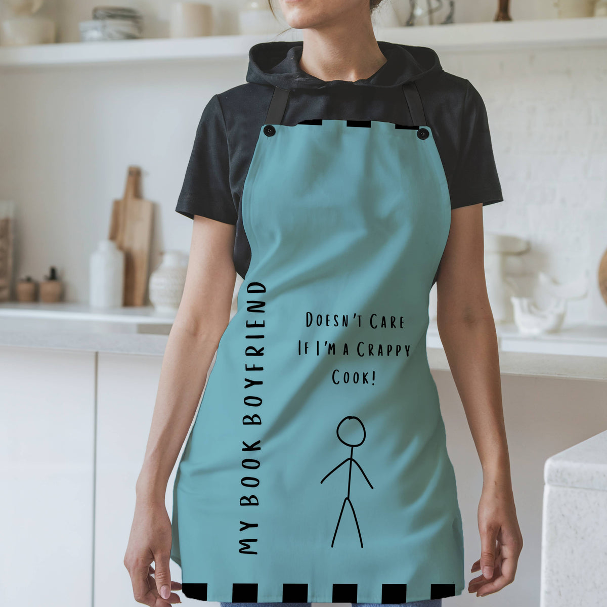 A woman wearing a teal apron with black straps stands in a modern kitchen. The apron features the phrase, "My Book Boyfriend" written vertically on the left side and "Doesn't Care If I'm a Crappy Cook!" in the center. A simple stick figure illustration is below the text. The apron has black square accents along the top and bottom edges. The background includes white shelves, cutting boards, and kitchen items.
