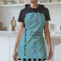 A woman wearing a teal apron with black straps stands in a modern kitchen. The apron features the phrase, "My Book Boyfriend" written vertically on the left side and "Doesn't Care If I'm a Crappy Cook!" in the center. A simple stick figure illustration is below the text. The apron has black square accents along the top and bottom edges. The background includes white shelves, cutting boards, and kitchen items.