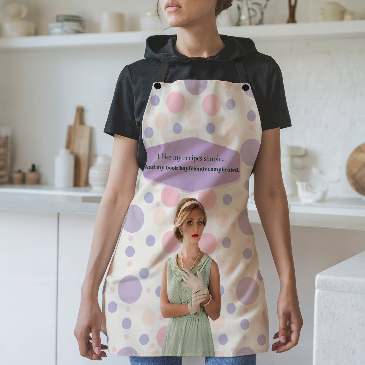 Woman in kitchen wearing white apron with pink and purple polka dots and black straps, vintage-style woman in mint green dress with pearls and gloves, text reads “I like my recipes simple and my book boyfriends complicated” inside purple speech bubble.