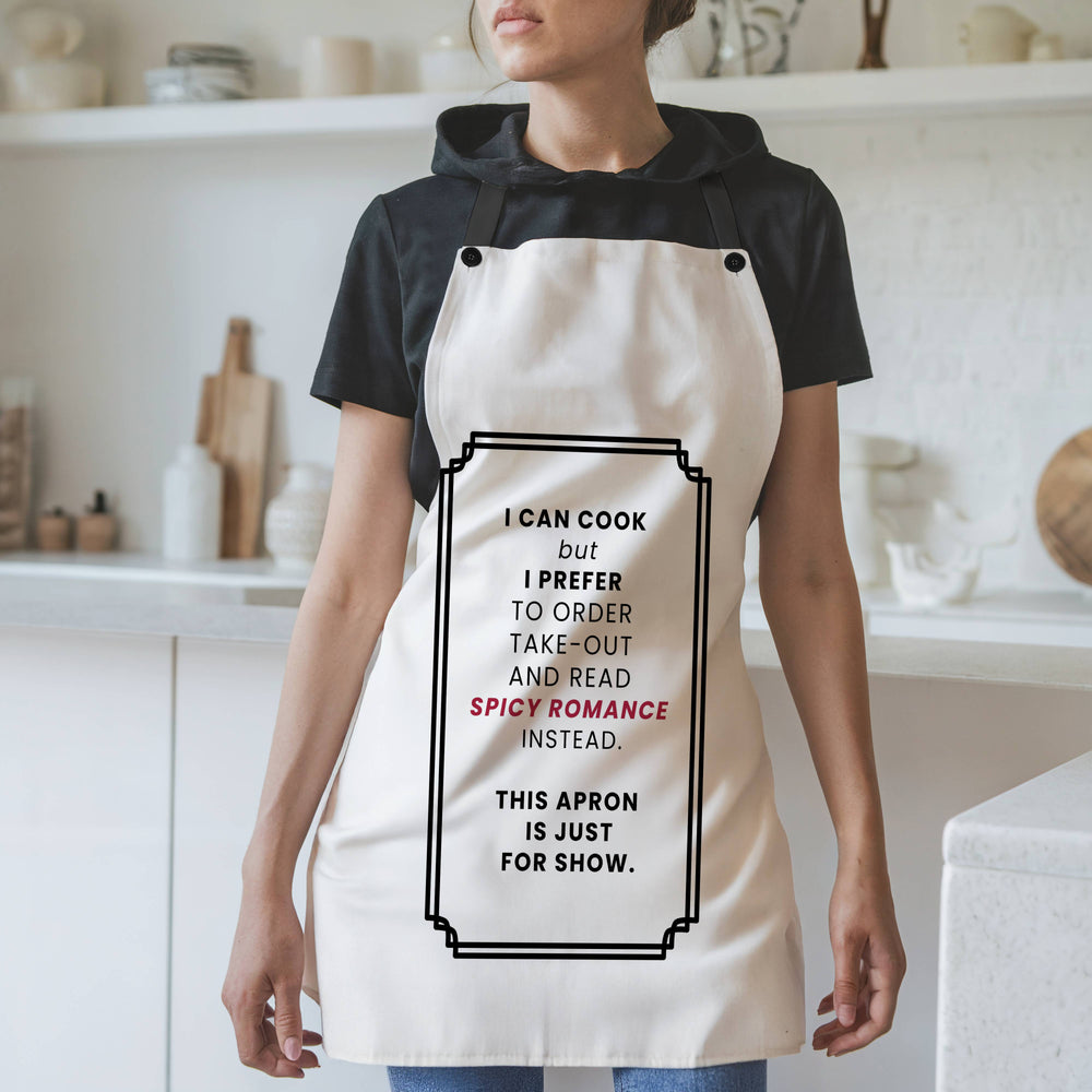 A woman wearing a white kitchen apron with a humorous black text graphic reading I can Cook but I prefer to order take-out and read spicy romance instead. this apron is just for show. It is styled over a black t-shirt in a bright, modern kitchen setting.