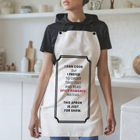 A woman wearing a white kitchen apron with a humorous black text graphic reading I can Cook but I prefer to order take-out and read spicy romance instead. this apron is just for show. It is styled over a black t-shirt in a bright, modern kitchen setting.