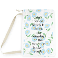 This design features a repeating pattern of light blue daisy-style flowers with yellow centers and green leaves on a white background. In the center, black serif text reads: “Can’t decide which is dirtier: my laundry or the romance books I read?” The floral pattern gives the design a soft, feminine touch, while the text delivers a cheeky, flirtatious message. The overall look is clean and pretty with a spicy twist for romance readers. There is a drawstring enclosure and a large canvas carry strap. 