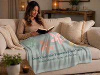 A woman sits on a couch reading a book with a sweatshirt  blaqnket over her lap.The blanket has a muted sage green  and light gray striped background with four stylized flowers in light saget green, coral pink, peach, and yellow, along with the text 'Flowers on the Outside. Morally Gray On The Inside.