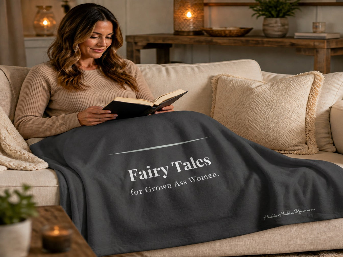 Woman reading on a sofa while wrapped in a dark gray sweatshirt blanket that reads "Fairy Tales for Grown Ass Women." Cozy blanket for romance readers who love book boyfriends and happy endings.