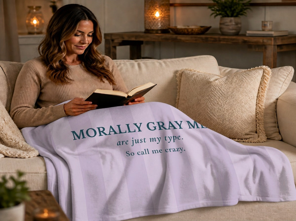 A woman is wrapped up in a  lavender and light gray striped blanket in a living room setting. The blanket features the text 'MORALLY GRAY MEN are just my type. So call me crazy.