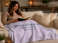 A woman is wrapped up in a  lavender and light gray striped blanket in a living room setting. The blanket features the text 'MORALLY GRAY MEN are just my type. So call me crazy.