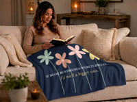 A woman sits on a beige sofa reading a book, wrapped in a navy blue sweatshirt blanket printed with large pastel flowers and the phrase, 'IF BOOK BOYFRIENDS WERE FLOWERS. I'd need a bigger vase