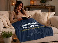 A woman sits on a beige sofa reading a book, wrapped in a navy blue blanket with white and gold text that reads, 'I LEARNED HOCKE THE HARD WAY. ONE BOOK BOY-FRIEND AT A TIME.