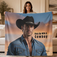 A woman holds up a large sweatshirt blanket featuring a printed portrait of a cowboy in a black hat and denim shirt against a sunset sky, with the text 'Cozy Up With A Cowboy' on a beige living room background.