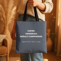A person in a beige sweater and jeans holds a dark grey tote bag with black handles. The bag features cream text that reads: 'CURSED. Dangerous. Morally Compromised. Apparently I have a type.